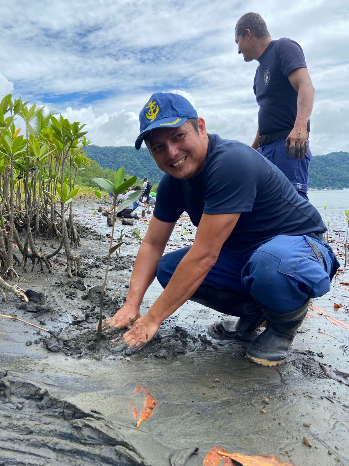 A young man kneels at the water's edge, planting a mangrove tree.