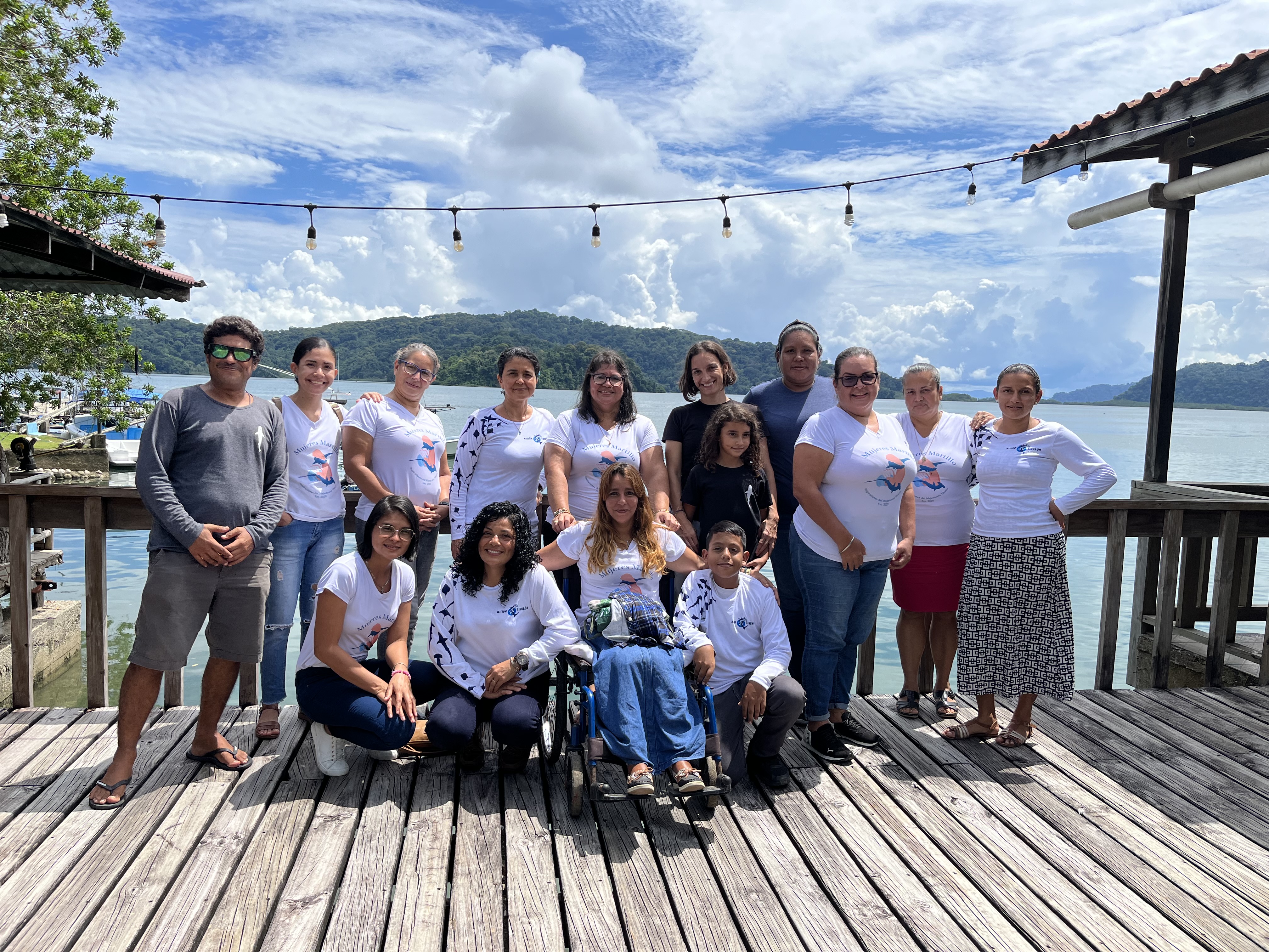 A group of 15 people pose for a photo in two rows on a veranda with water in the background.