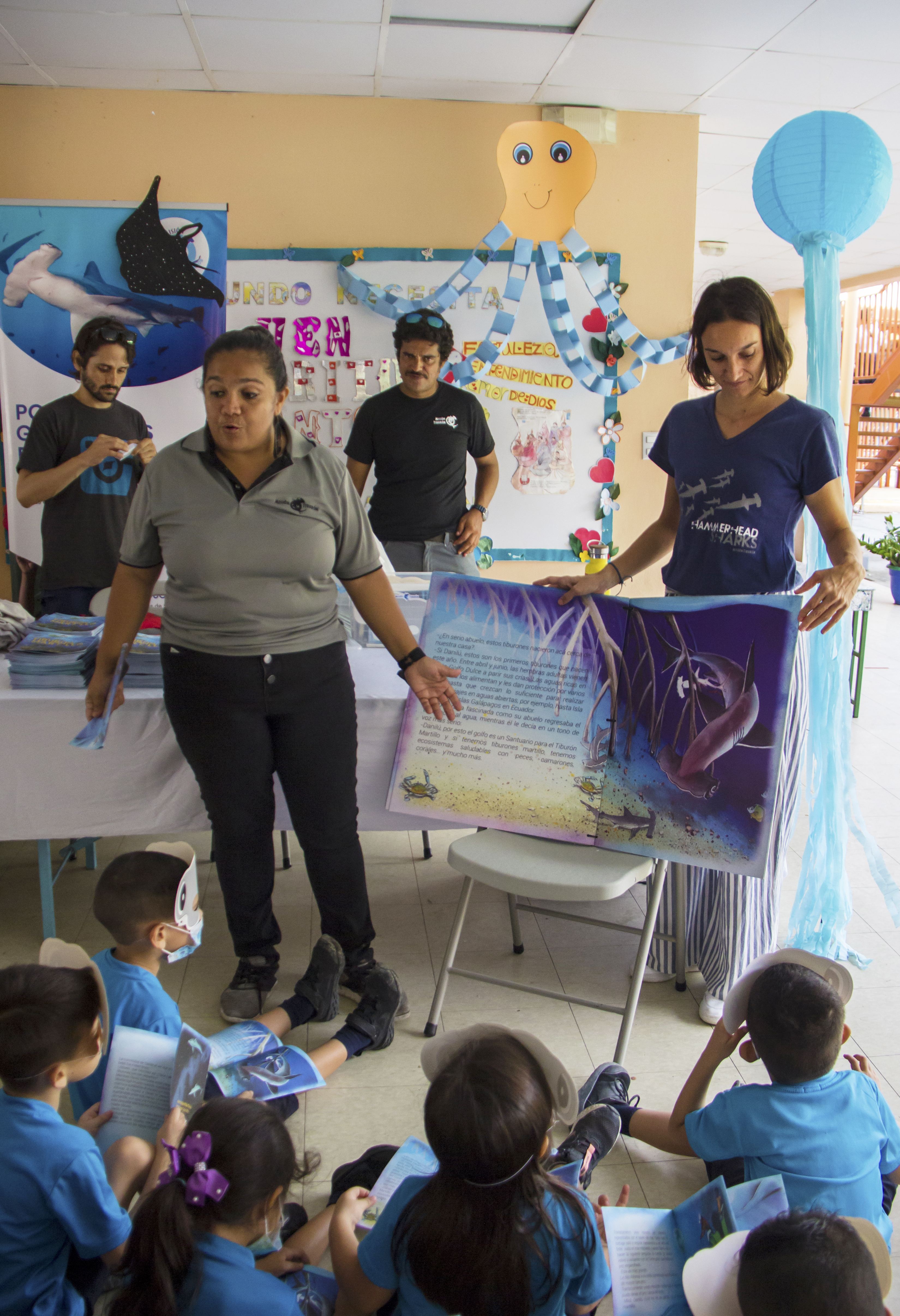 A classroom setting with two female teachers standing in front of a group of seated young students.
