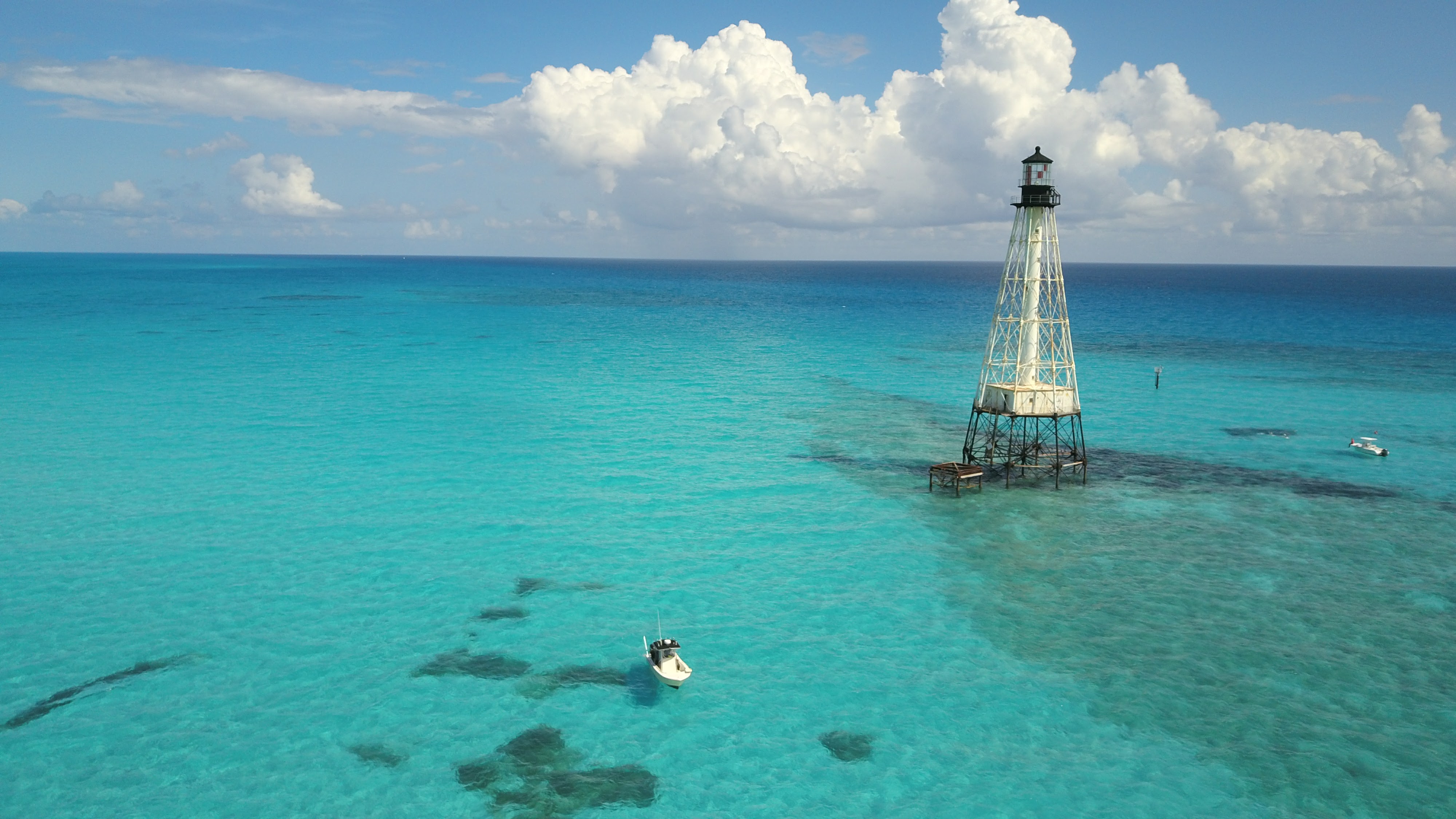 A solitary lighthouse stands on tall metal stilts over turquoise waters, with a small boat nearby and scattered clouds in the sky.