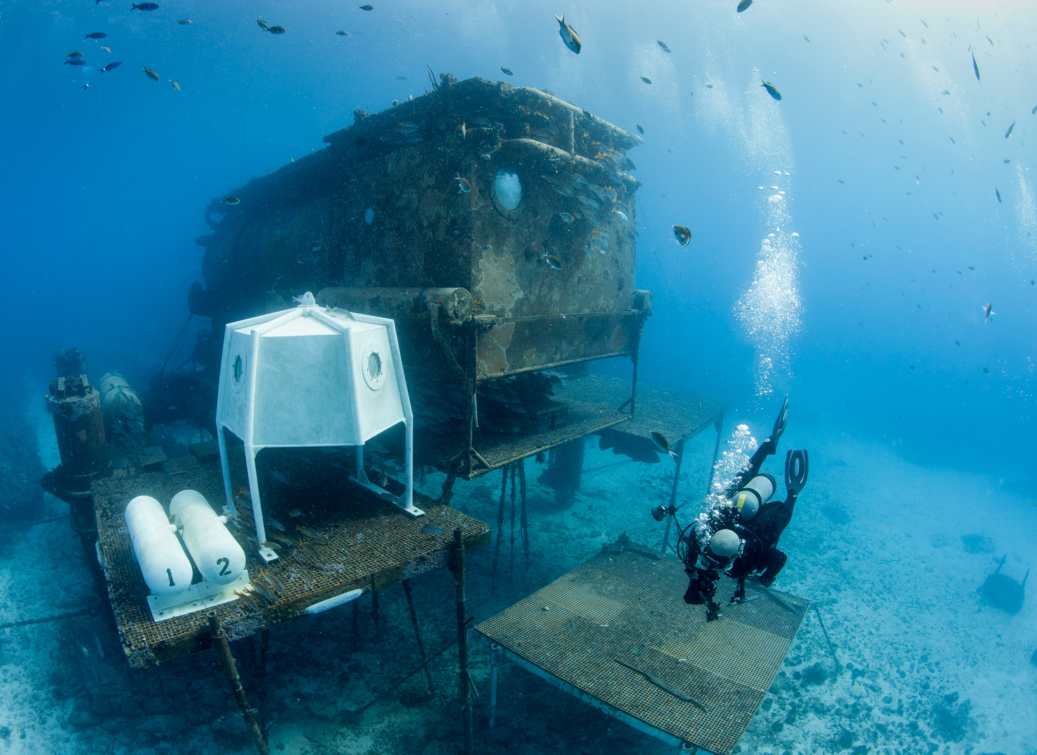 A scuba diver explores an underwater habitat structure, resting on metal platforms. The habitat appears aged and covered in marine growth. Small fish swim around, and the divers' bubbles rise to the surface in the clear blue water.