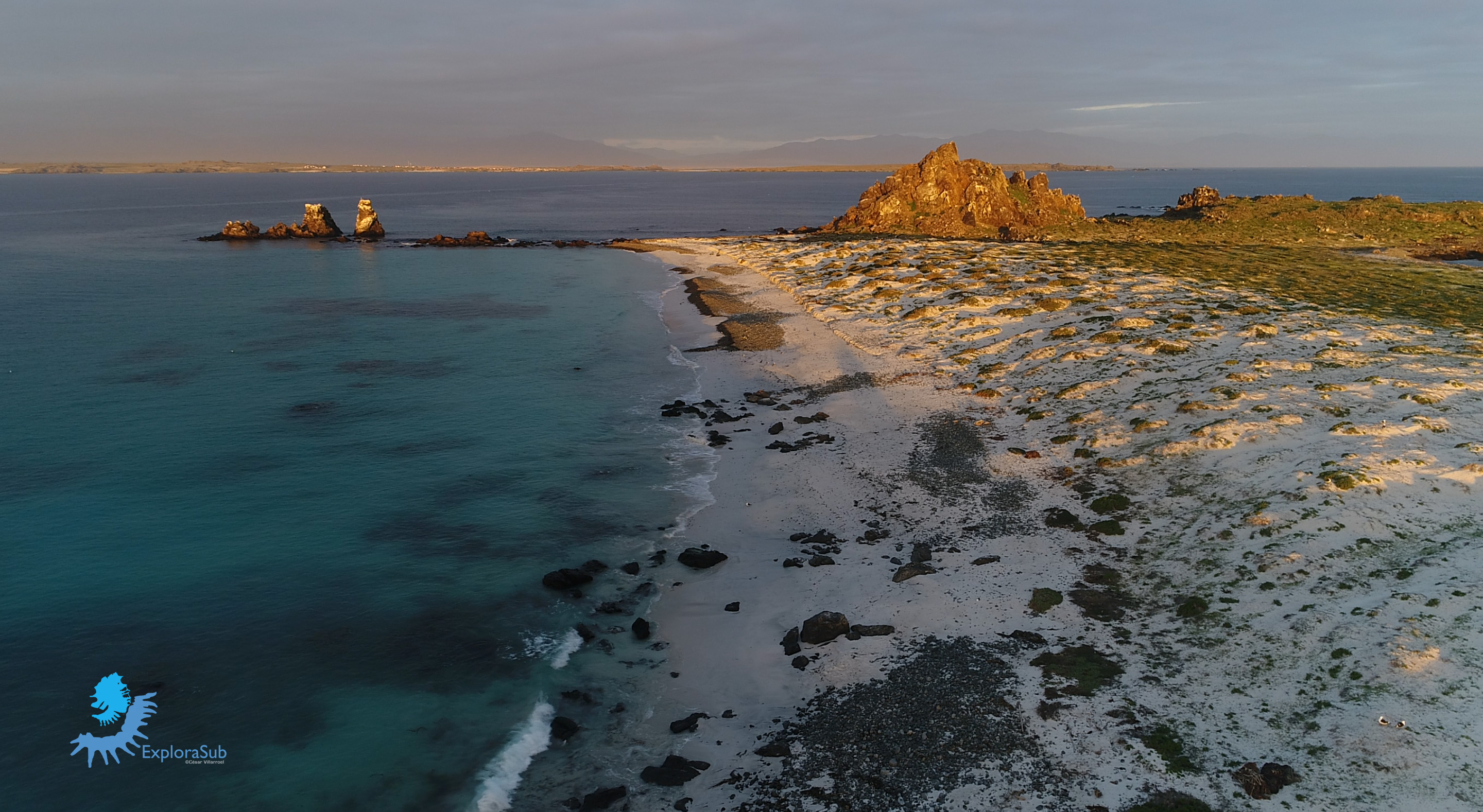 Aerial view of a rocky coastline with blue water and sandy beaches, featuring a prominent rock formation in the background.