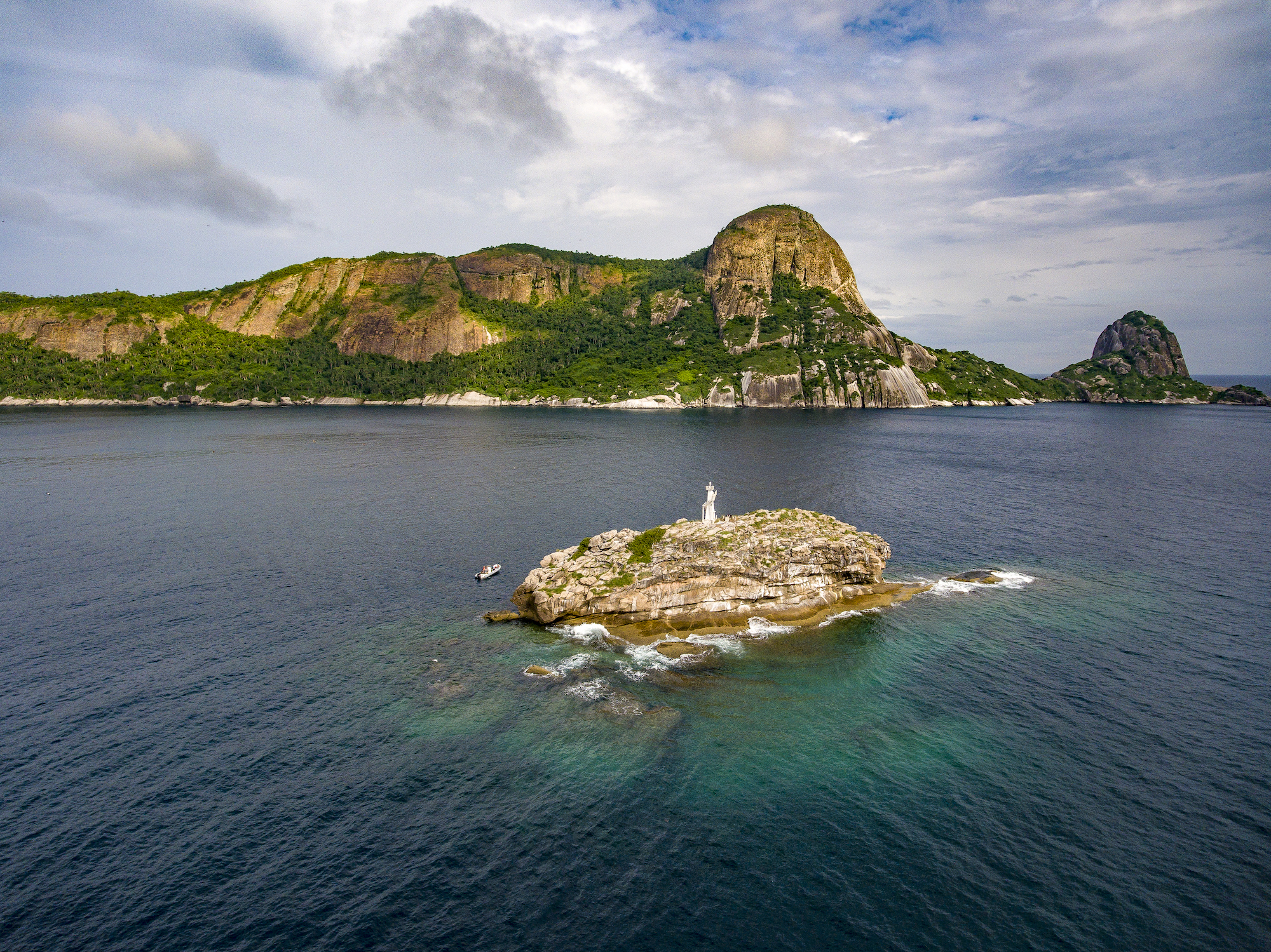 An aerial view of a small, rocky island with a white statue on it, surrounded by blue-green ocean water. In the background, green hills with rocky cliffs rise under a partly cloudy sky. A boat is near the island.