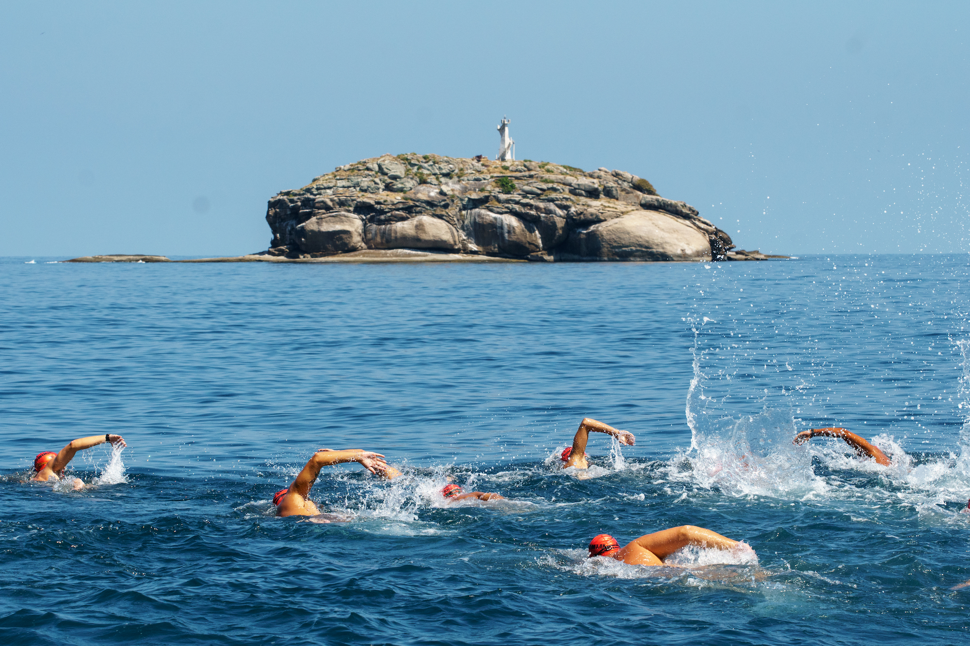 A group of swimmers wearing red swim caps are swimming in an open body of water towards a small rocky island with a white statue or monument on top. The sky is clear, and the water is calm except for the splashing caused by the swimmers.