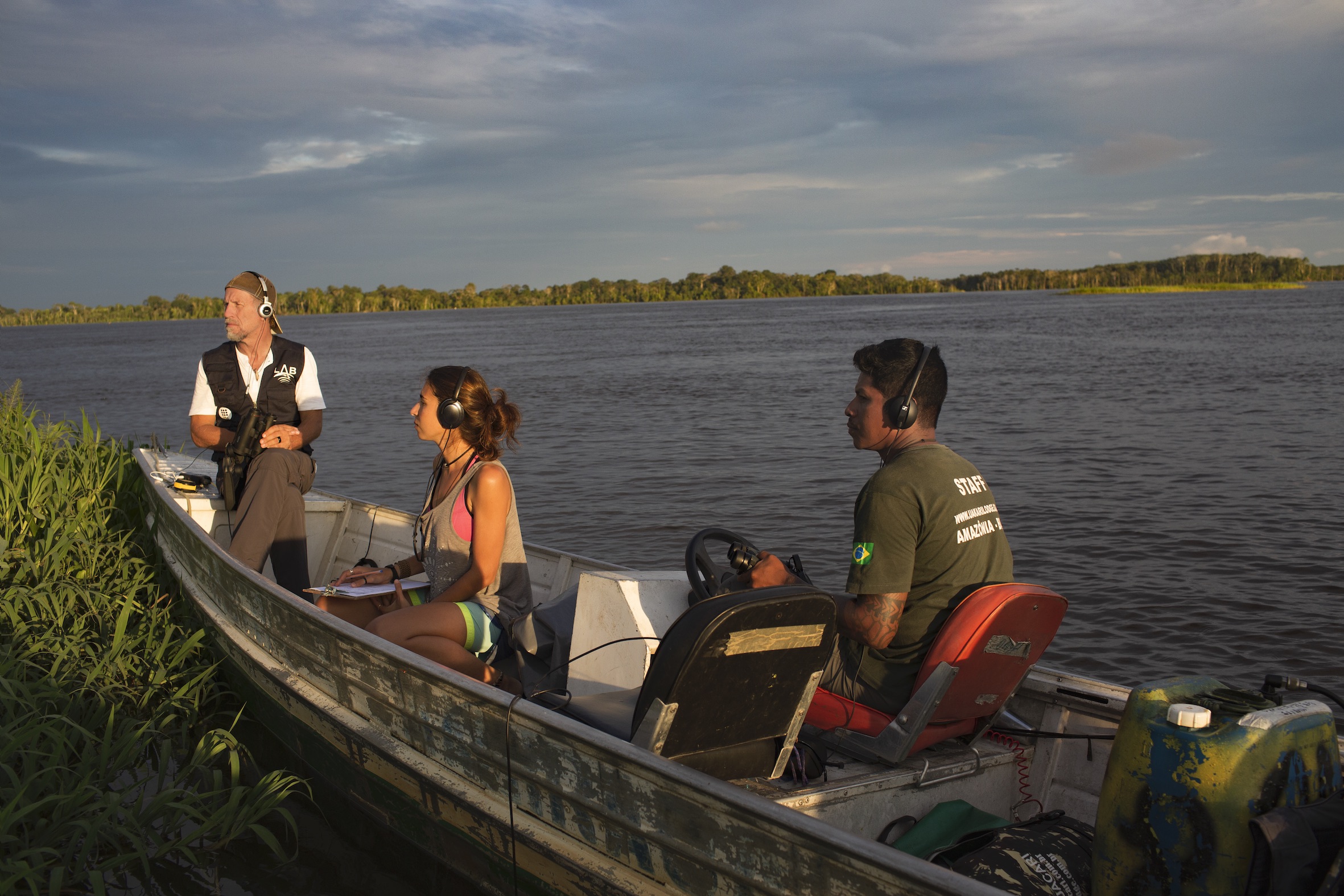 Three individuals sit in a small boat on a body of water surrounded by grassy banks. They all wear headphones and seem to be listening attentively. One man is steering the boat while the other two, a man and a woman, observe the surroundings and take notes.