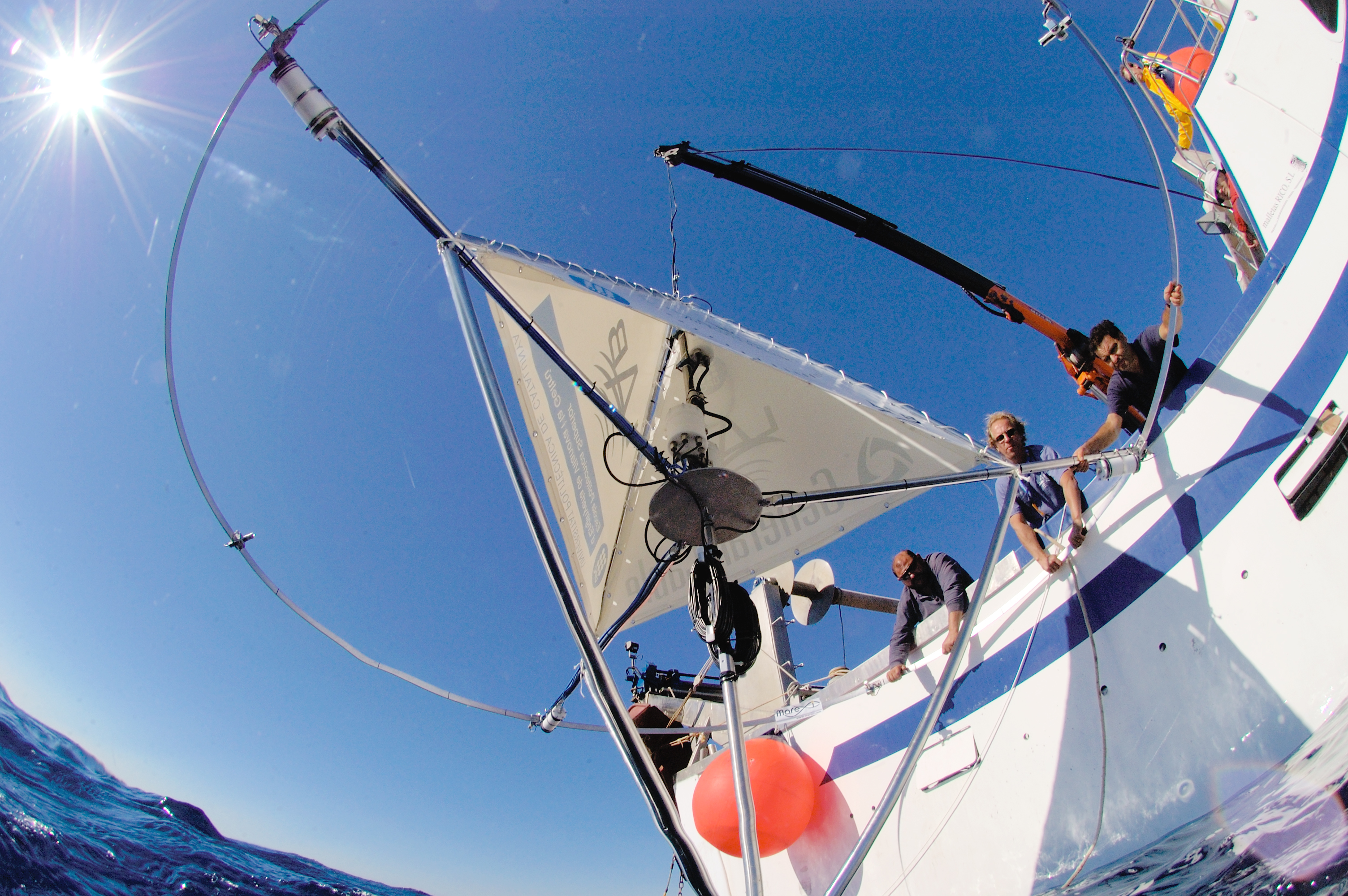 A broad-angle view of a sailing boat on the ocean. Several people are onboard, adjusting equipment and enjoying the sunny day. The sky is clear with the sun shining brightly overhead. The boat features sails and rigging, and a vibrant red buoy is attached to its side.