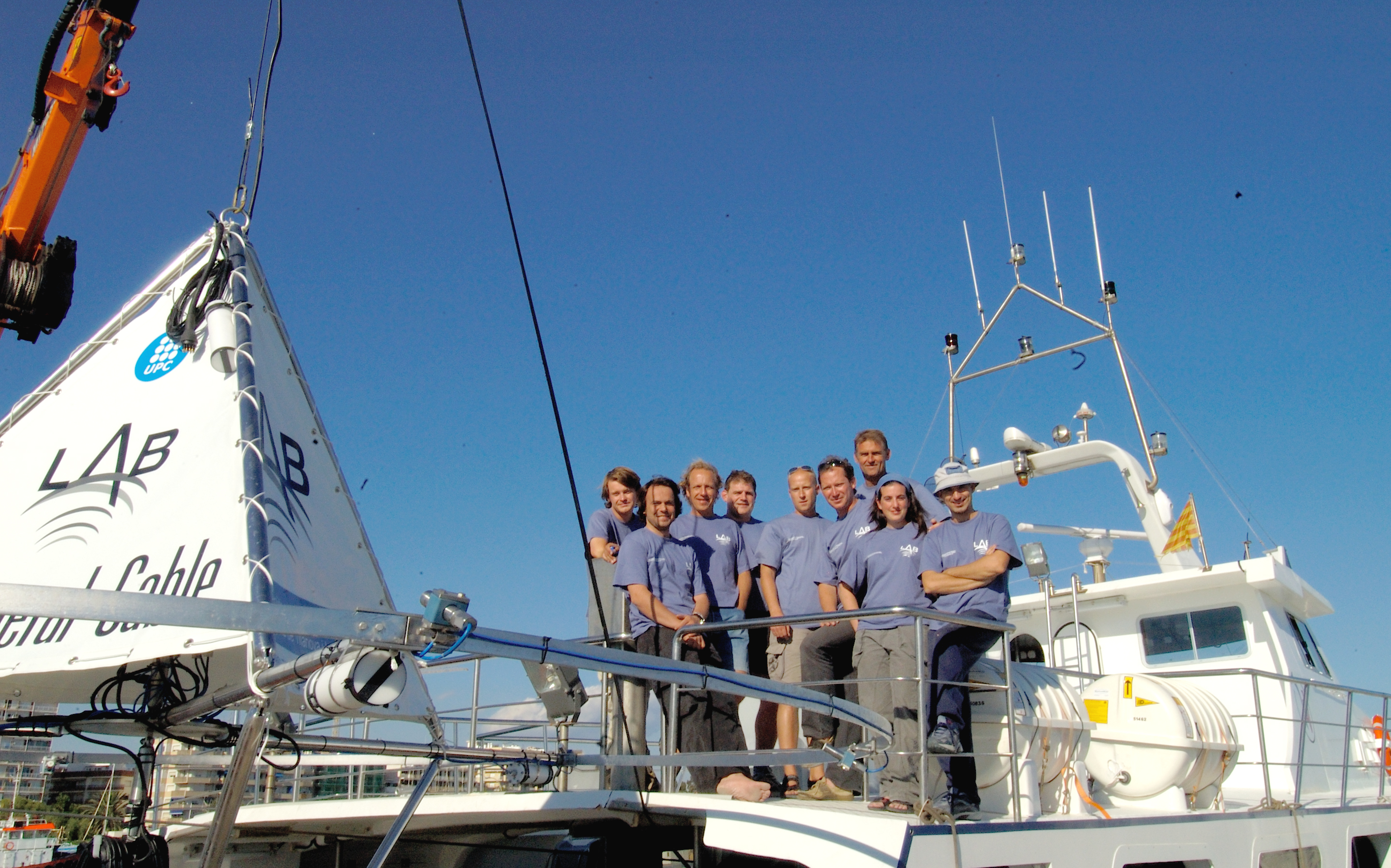 A group of eight people, wearing blue shirts, standing together on the deck of a boat with various equipment. The boat has a white sail with the word "LAB" printed on it. The sky is clear and blue, and the background includes some buildings and a dock.