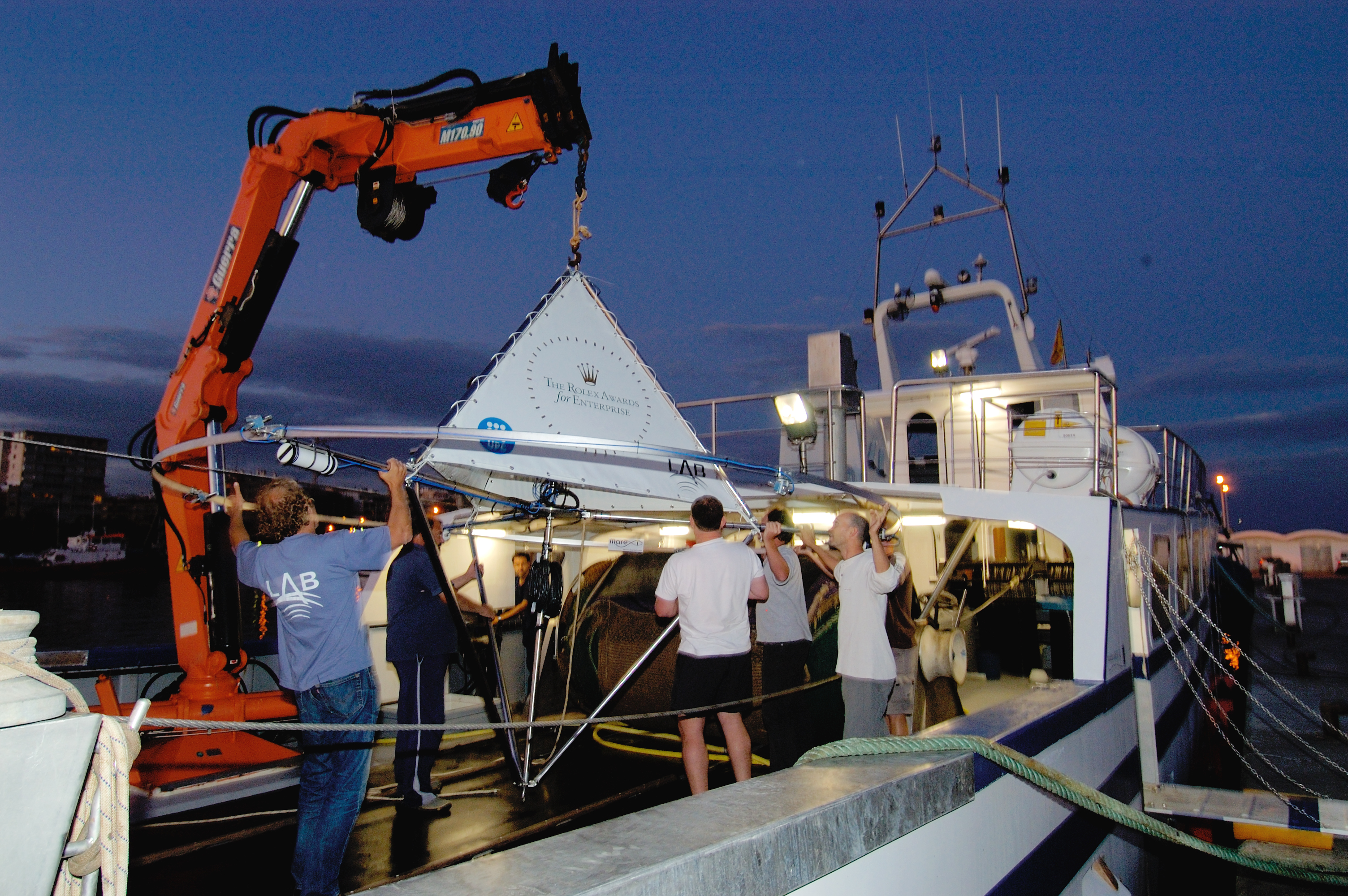 A group of people on a boat use a mechanical crane to lift a triangular piece of equipment from a boat at dusk. The equipment features a metal framework and a white triangular section with text on it. The boat has a control station illuminated by lights.