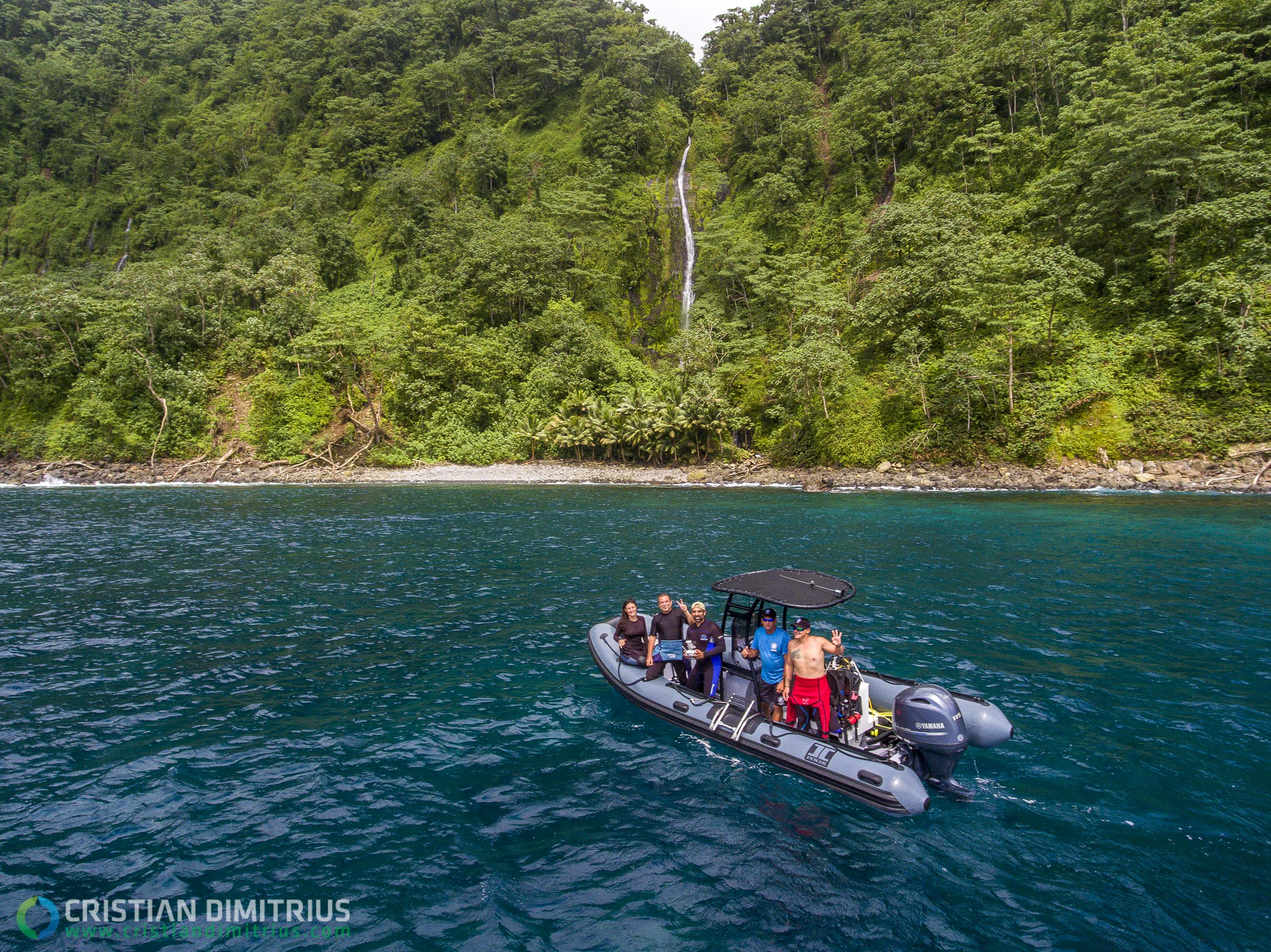 A small inflatable boat with a canopy sails just off the shore of a white beach with dense green vegetation. 