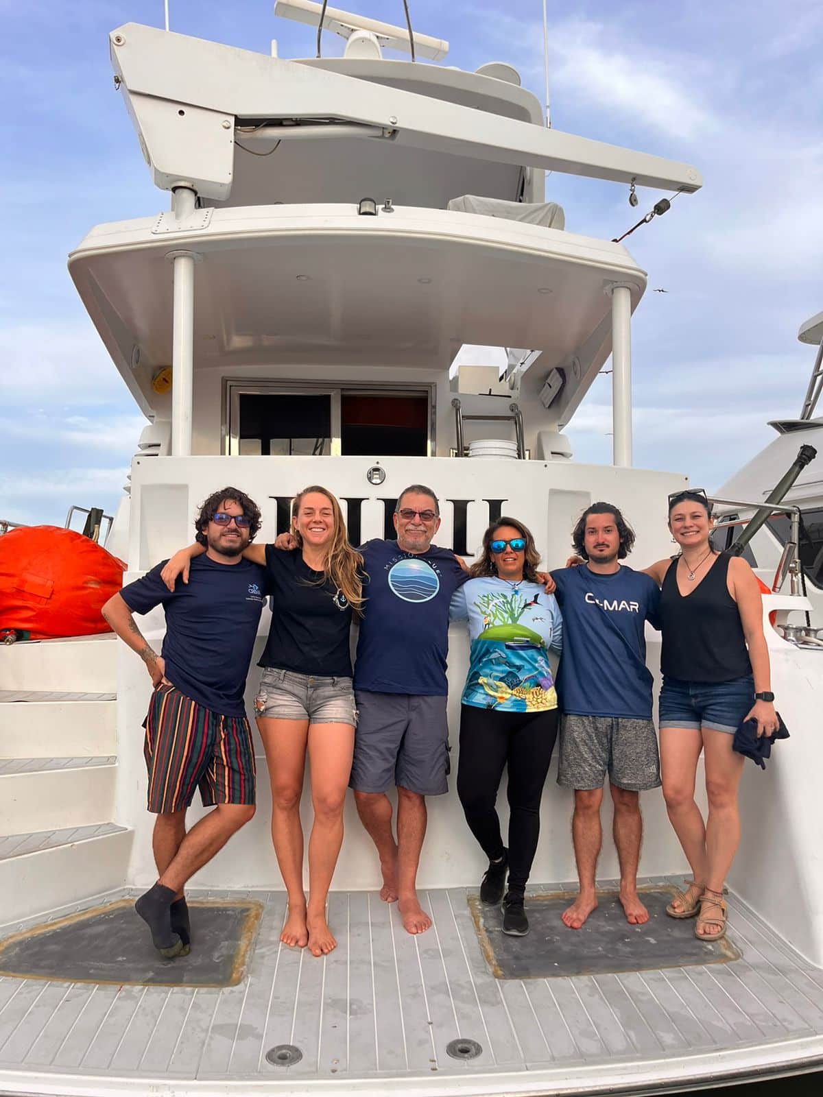 A group of six people stand on the back of a boat. 